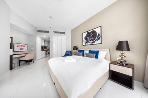 A modern hotel room with a large bed, a desk, a chair, a bedside table with a lamp, and a painting on the wall. - Stayshort - Cozy Studio in Damac Hills 1 for 2 With Pool Access (Dubai)