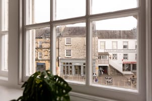 Blick durch ein Fenster mit einer Pflanze im Vordergrund, auf eine Straße mit Gebäuden, darunter ein Pub und ein Herrenhaus.