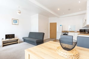Un apartamento moderno con un sofá gris, una mesa de comedor de madera, una cocina con armarios blancos y un televisor pequeño sobre un mueble de madera.