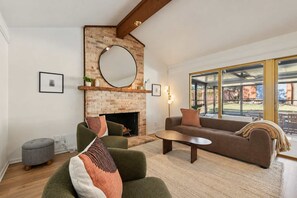 Ein Wohnzimmer mit Kamin aus Ziegelsteinen, einem runden Spiegel, einer Couch, einem Couchtisch und Blick durch große Fenster.