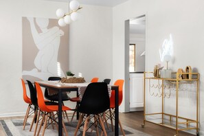Ein Essbereich mit einem Holztisch, orangefarbenen und schwarzen Stühlen, einem gewebten Korb und einem goldrahmenartigen Beistelltisch mit Gläsern.