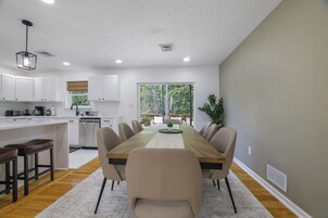 Cozinha moderna com armários brancos, área de jantar com mesa e cadeiras de madeira, e vista para o verde através de grandes janelas.