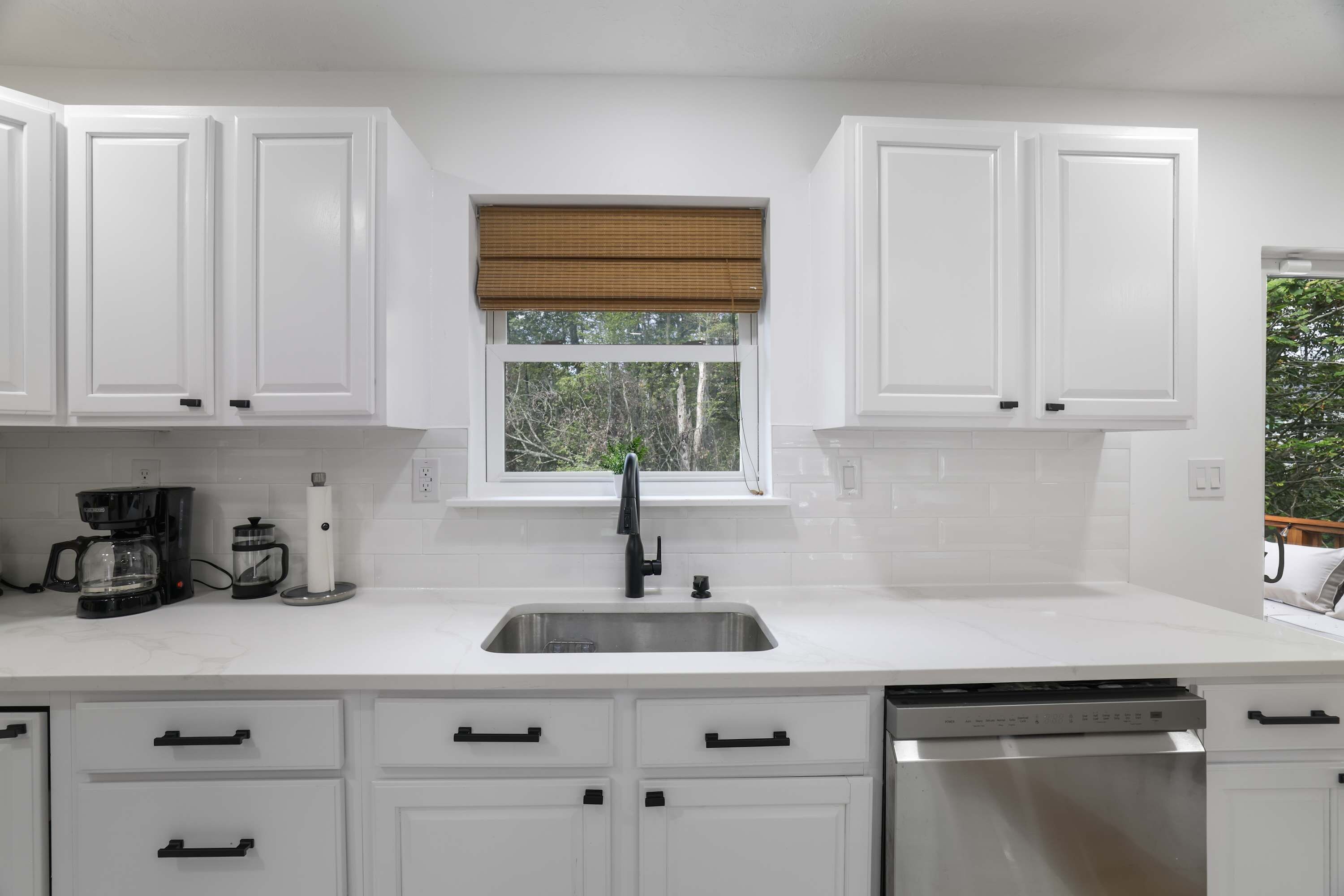 A kitchen with white cabinets, a stainless steel sink, a coffee maker, and a window with a blind.