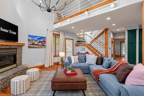 A spacious living room with a fireplace, a large sofa, a coffee table, and a staircase leading to an upper level. - Helen Haus Golf Sim Hot Tub Theater Retreat (Helen)