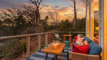 A balcony with a wooden table, a blue sofa with red cushions, and a view of trees and a sunset.