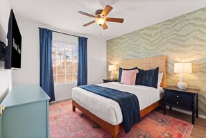 Ein Hotelzimmer mit Bett, Nachttischen, Deckenventilator, Fenster mit Vorhängen und einer Wand mit Muster.