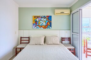 A bedroom with a bed, two chairs, a colorful abstract painting on the wall, and a window with blinds. - Apartment in Condominium Itagua Viewpoint - Between Praia Grande and Tenório - 10 min walk (Ubatuba)