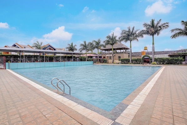 A swimming pool with a metal ladder, surrounded by palm trees and a covered area with a thatched roof. - Townhouse Oak Rising Water Park Dharwad (Dharwad)