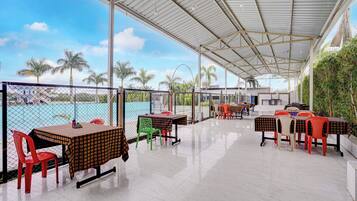 A covered outdoor seating area with tables and chairs, a pool in the background, and palm trees.