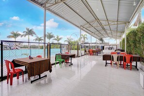 A covered outdoor seating area with tables and chairs, a pool in the background, and palm trees. - Townhouse Oak Rising Water Park Dharwad (Dharwad)