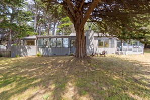 Ein zweigeschossiges Haus mit einem großen Baum davor, einem eingezäunten Garten und einer Terrasse mit Sitzgelegenheiten.