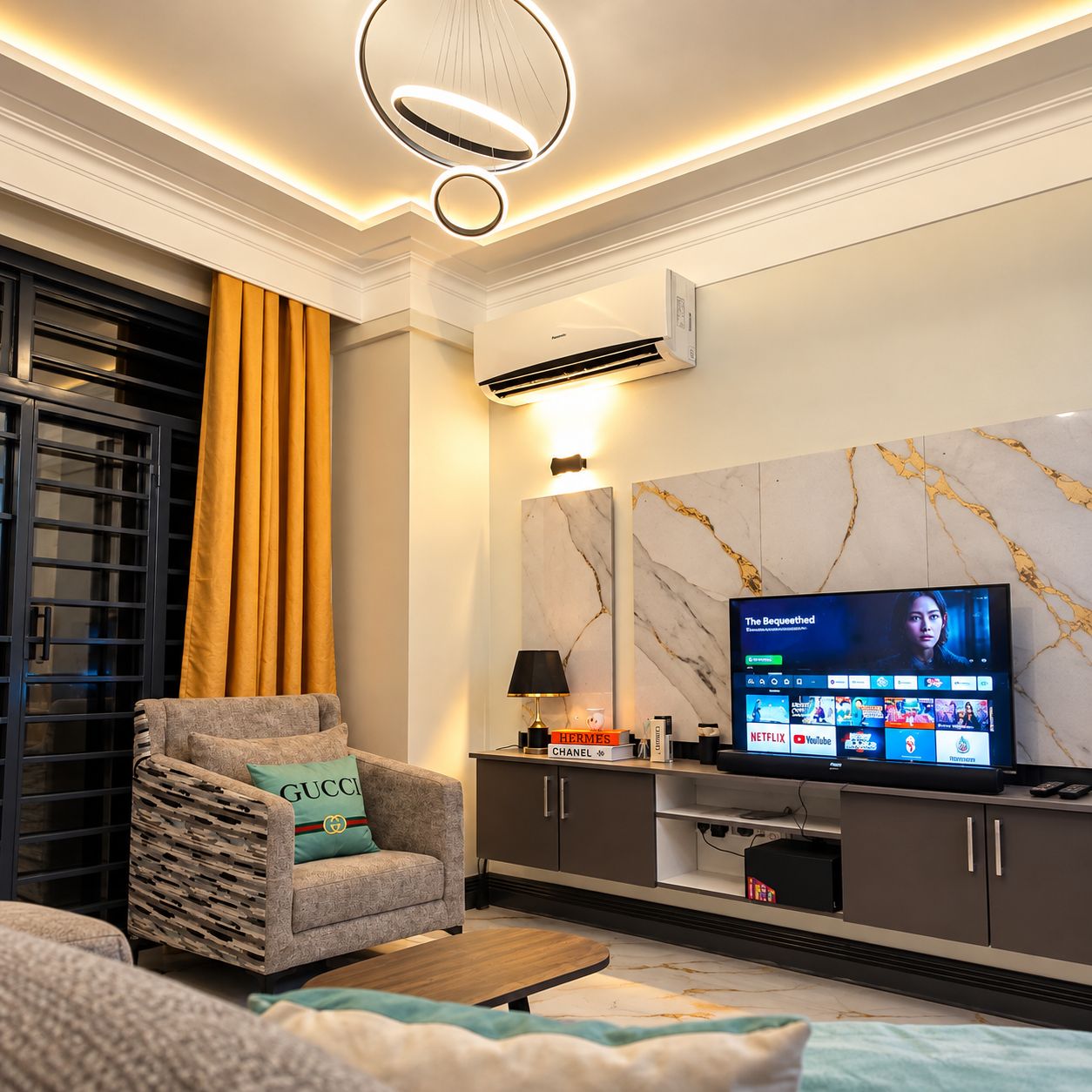 Una sala de estar moderna con un televisor de pantalla plana, un sofá con un cojín Gucci, una pared con diseño de mármol y un ventanal con cortinas.