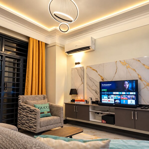 Ein modernes Wohnzimmer mit einem Flachbildfernseher, einem Sofa mit einem Gucci-Kissen, einer Wand mit Marmorstruktur und einem großen Fenster mit Vorhängen.