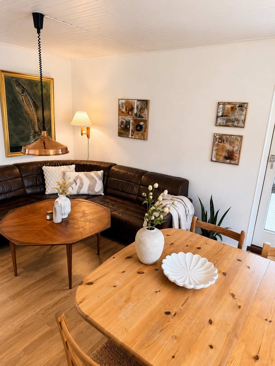 Un salon avec un canapé en cuir marron, une table basse en bois, une table à manger ornée d’un vase blanc et d’un coquillage, et une lampe fixée au mur.