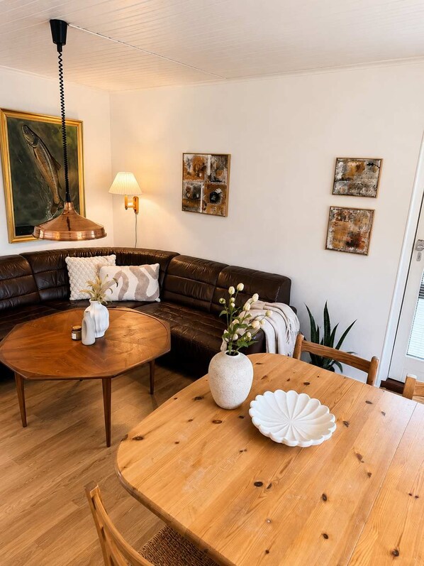 A living room with a brown leather sofa, a wooden coffee table, a dining table with a white vase and a shell, and a lamp on the wall.