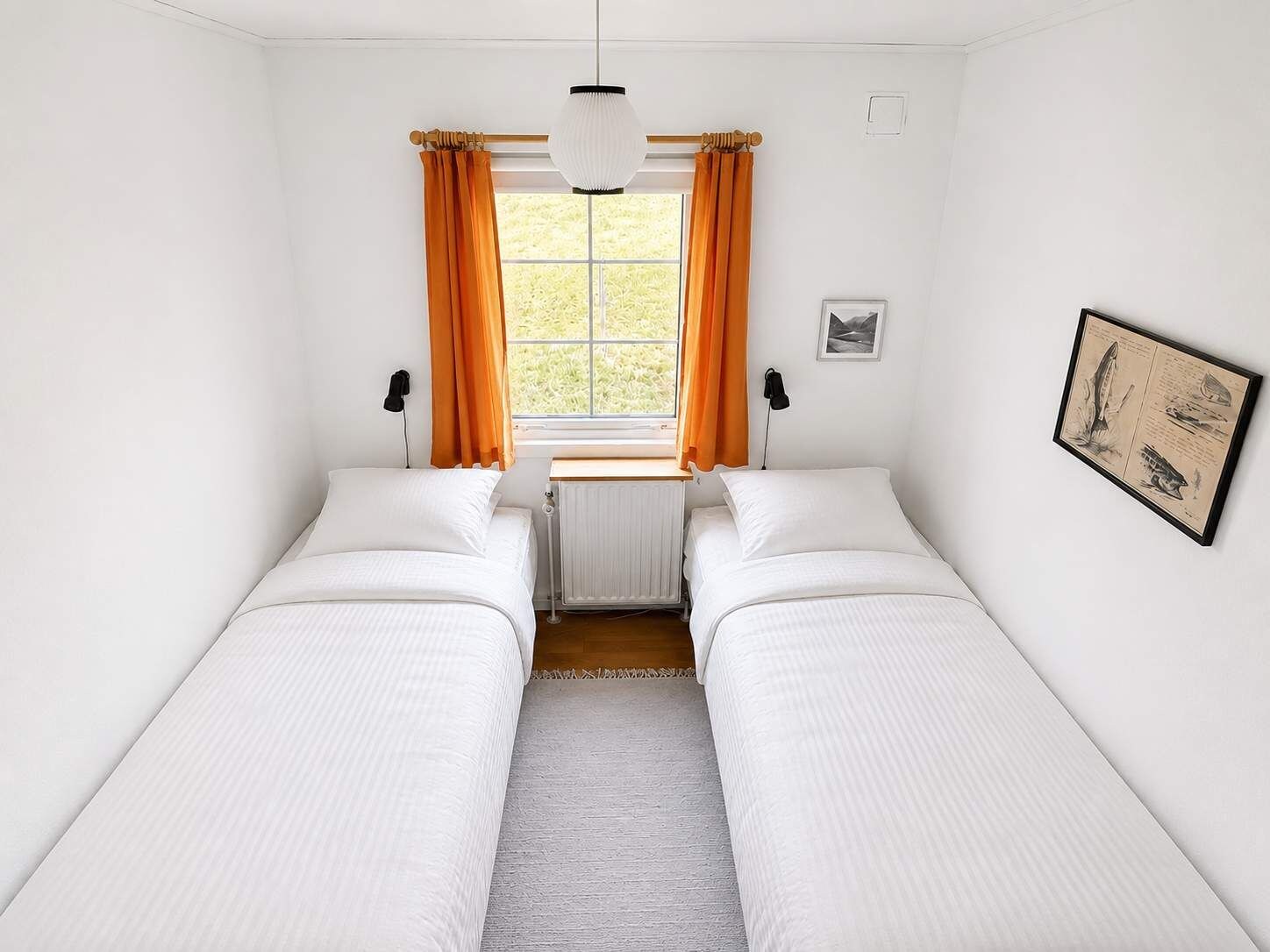 A room with two single beds, a radiator, a window with curtains, a hanging light fixture, and a framed picture on the wall.