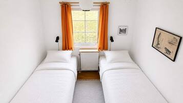 A room with two single beds, a radiator, a window with curtains, a hanging light fixture, and a framed picture on the wall.