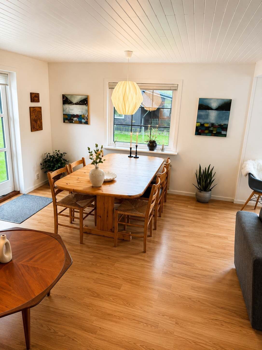 A dining area with a wooden table, chairs, a vase with flowers, and a hanging light fixture.
