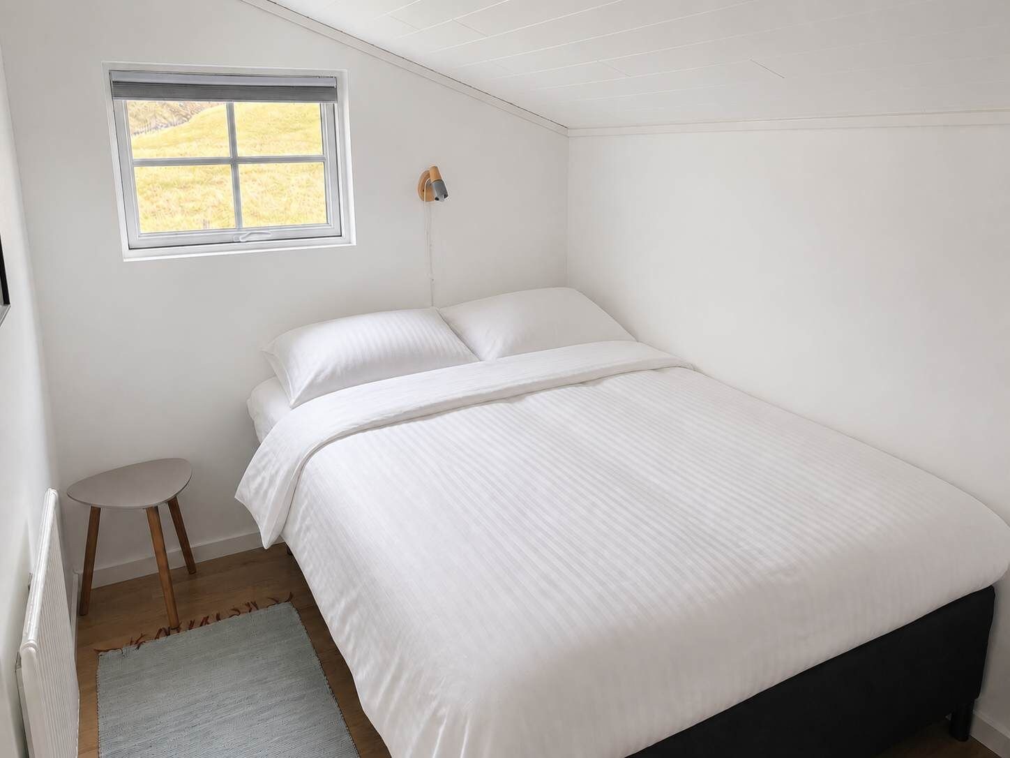 A bedroom with a bed, a small stool, a window, and a radiator.