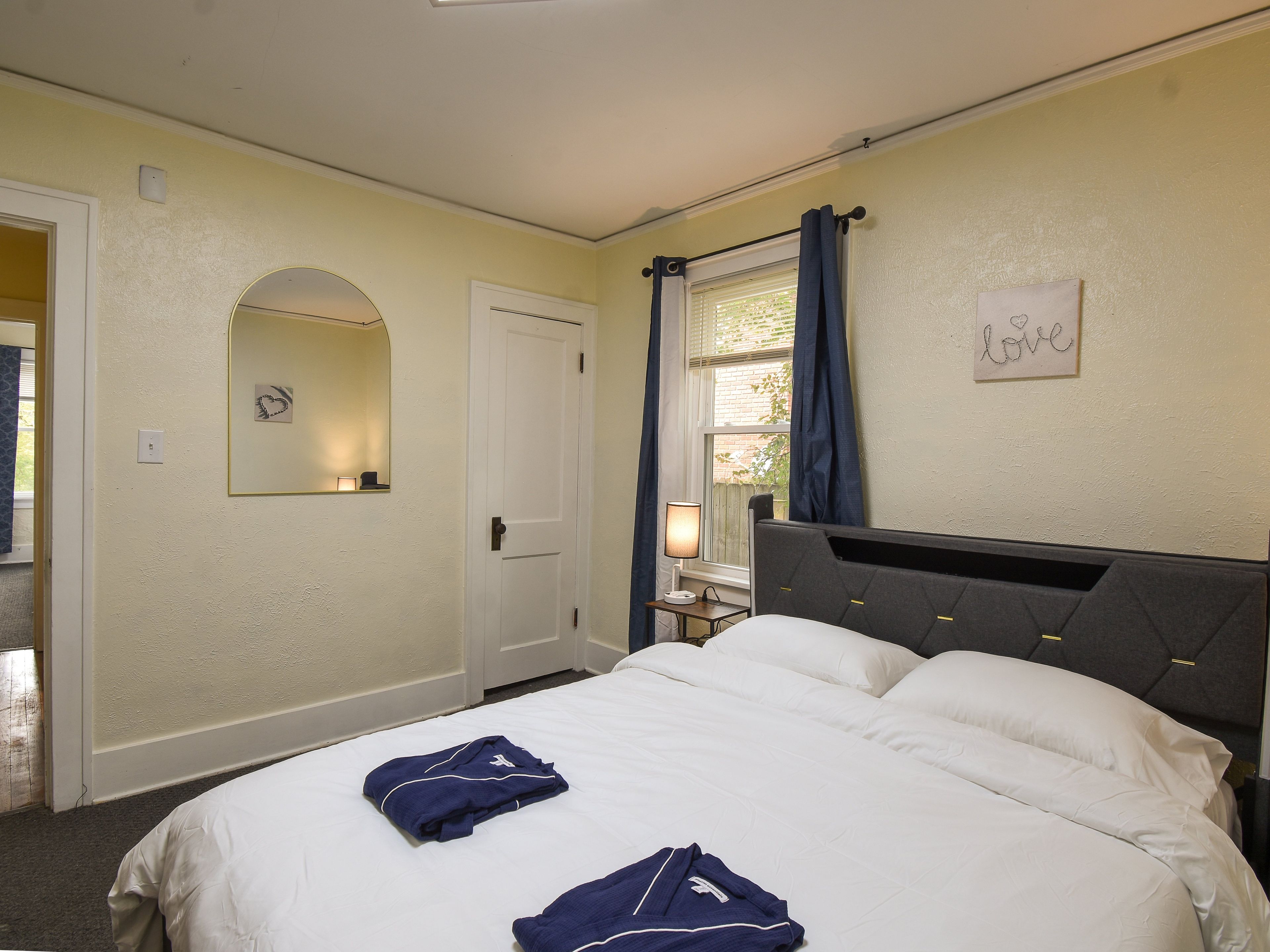 A bedroom with a bed, a bedside table with a lamp, a mirror, a door, and a window with curtains.