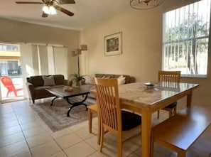 Ein Wohnzimmer mit einer Couch, einem Couchtisch und einem Esstisch mit Stühlen. Es gibt ein Fenster mit Jalousien und einen Deckenventilator.