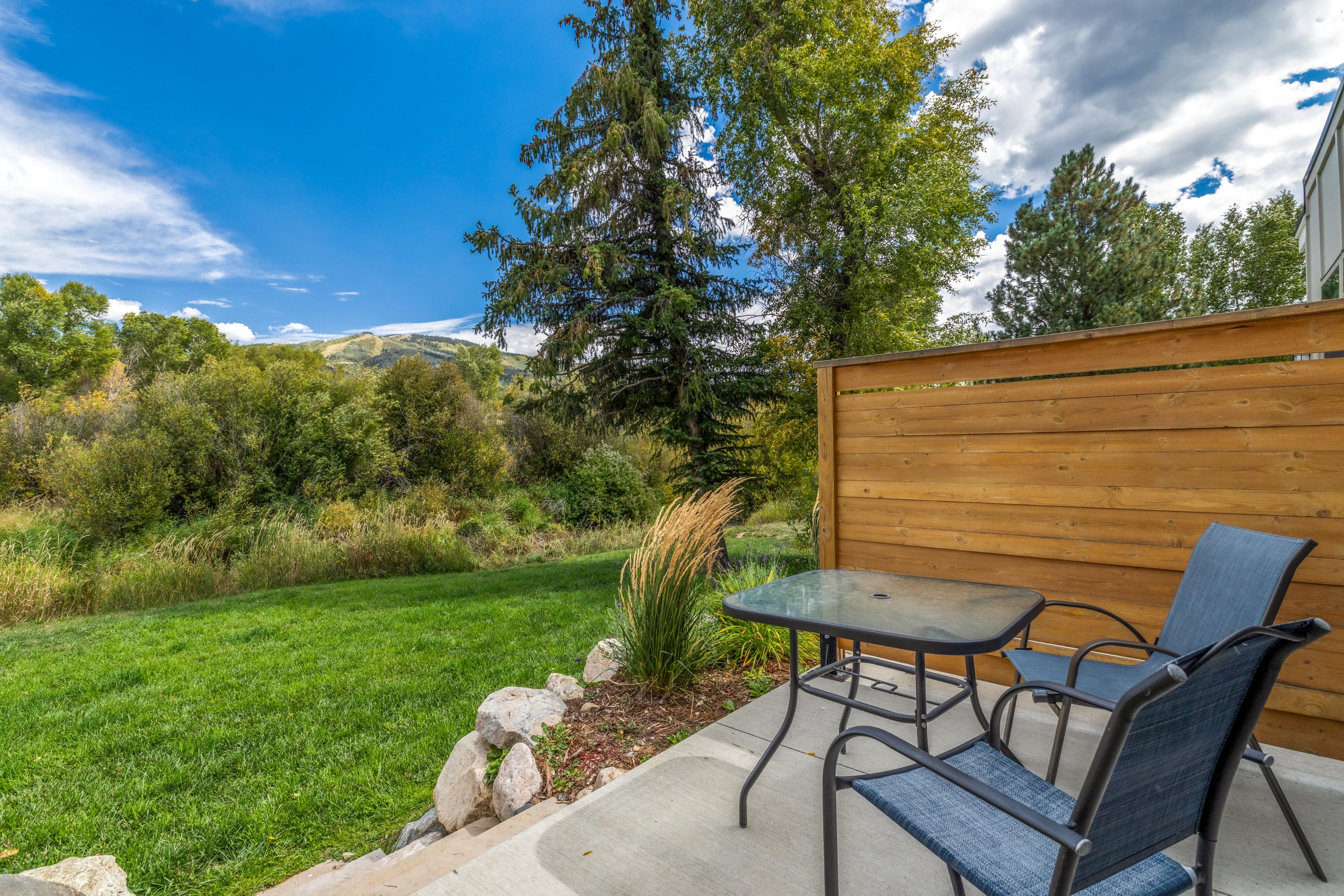 An outdoor patio with a table and chairs, surrounded by a wooden fence, with a view of a grassy area and trees.