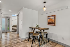 Un comedor con una mesa de madera y cuatro taburetes, un cuadro en la pared y una escalera al fondo.