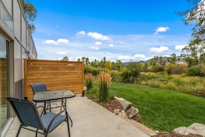 Un patio con una mesa y sillas, una cerca de madera y vistas a un paisaje con césped, árboles y colinas.
