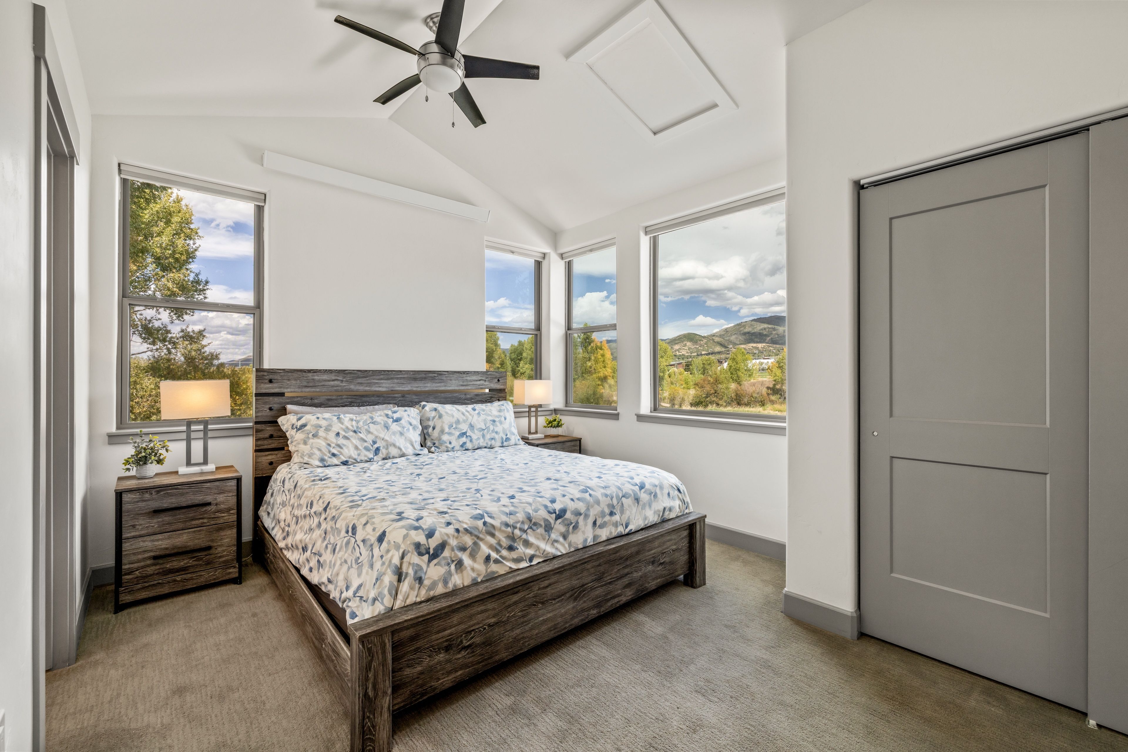 A bedroom with a bed, two bedside tables, a ceiling fan, and large windows offering a view of the outdoors.
