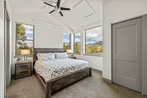 Un dormitorio con cama, dos mesitas de noche, ventilador de techo y ventanas amplias con vistas al exterior.
