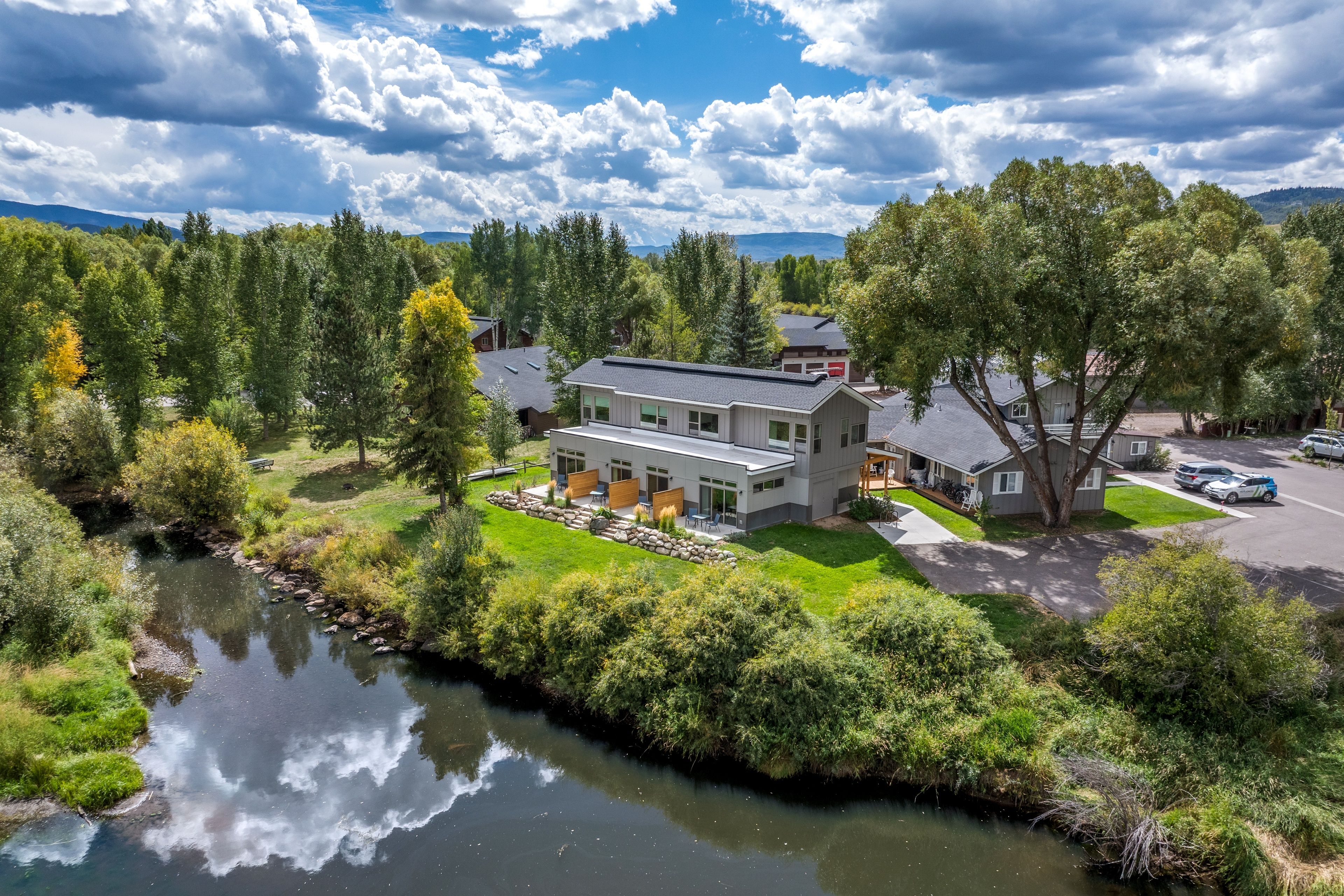 A modern house with a large patio area, a river in the foreground, and a mountainous backdrop.