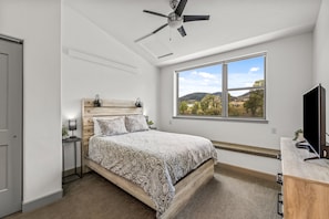 A bedroom with a bed, a ceiling fan, a window with a view, and a desk with a computer.