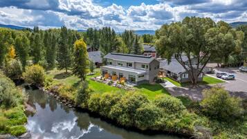 A modern house with a large patio area, a river in the foreground, and a mountainous backdrop.