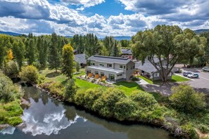 A modern house with a large patio area, a river in the foreground, and a mountainous backdrop.