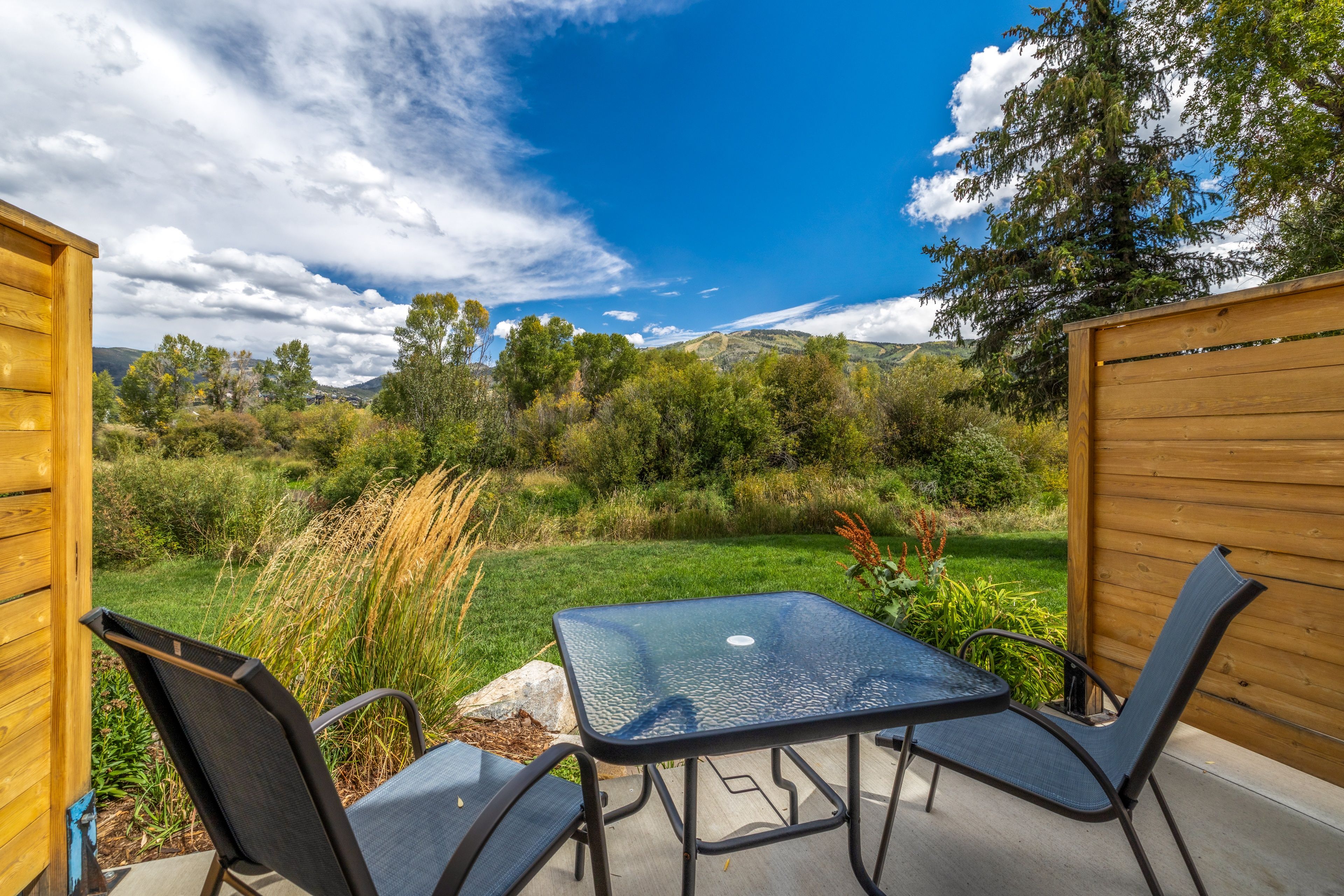 An outdoor patio with a glass table and two chairs, surrounded by a wooden fence, overlooking a grassy area with trees and hills.