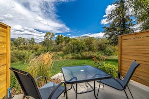 An outdoor patio with a glass table and two chairs, surrounded by a wooden fence, overlooking a grassy area with trees and hills.