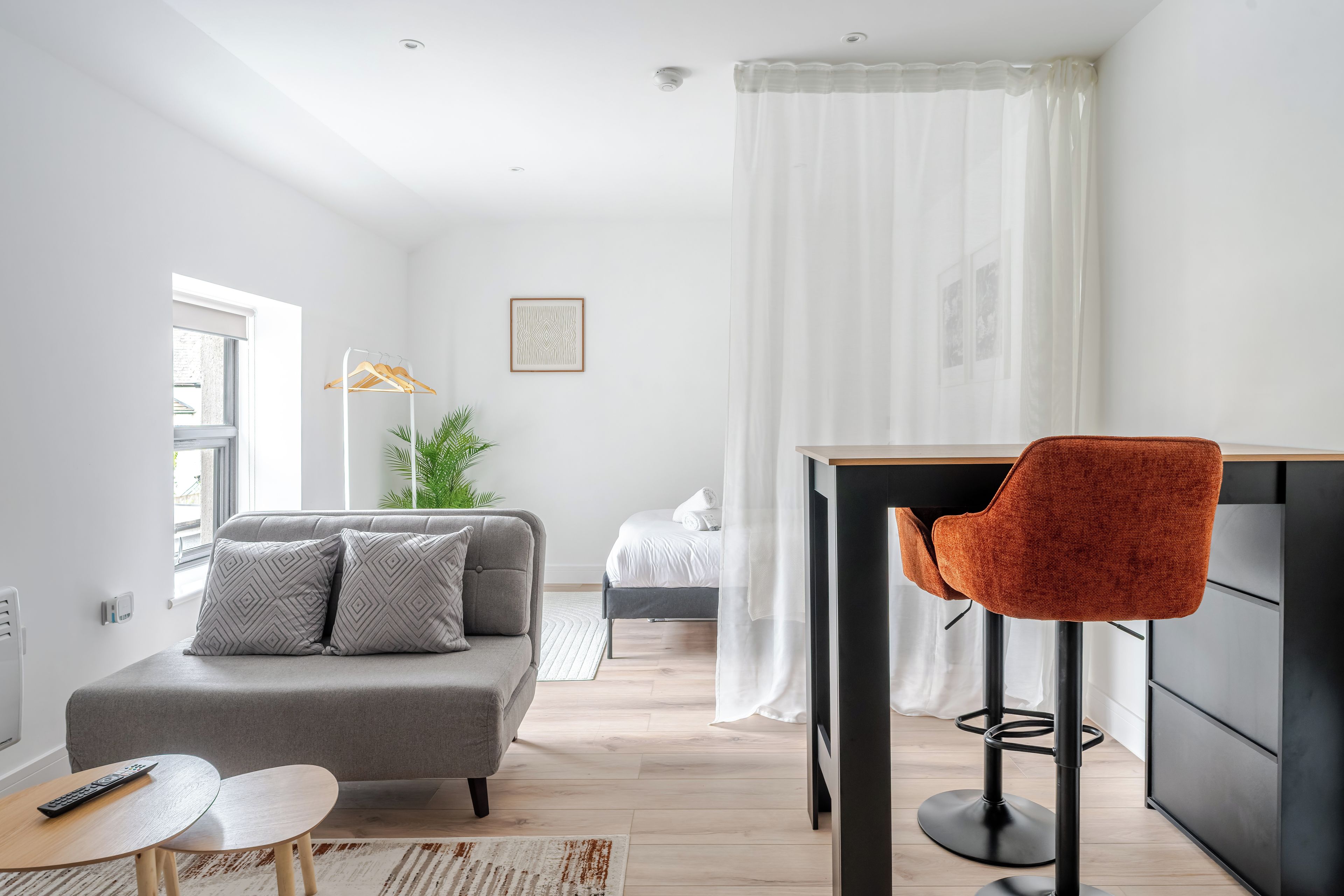 A modern living room with a grey sofa, a small round table, a red chair, and a bed in the background.