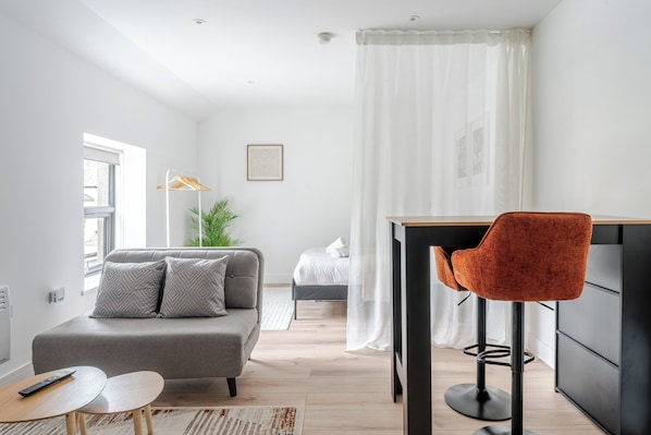 A modern living room with a grey sofa, a small round table, a red chair, and a bed in the background.