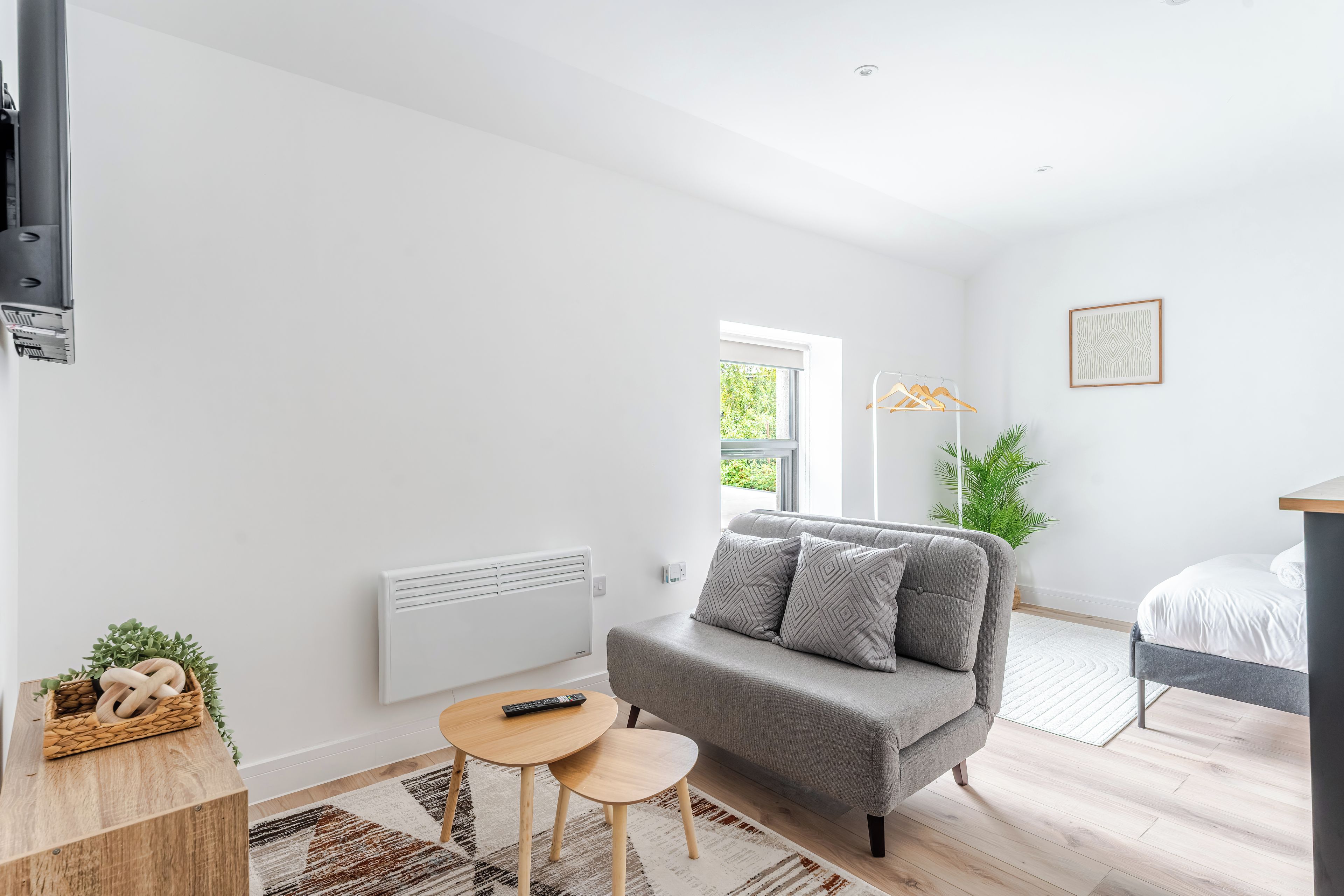 A modern, well-lit room with a grey sofa, a small round table, a radiator, a potted plant, and a bed with white bedding.