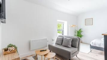Une pièce moderne et bien éclairée, avec un canapé gris, une petite table ronde, un radiateur, une plante en pot et un lit recouvert de linge de lit blanc.