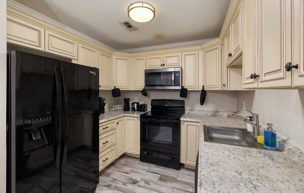 A kitchen with cream-colored cabinets, a black refrigerator, a microwave, an oven, and a sink with a marble countertop.