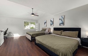 A bedroom with two beds, a ceiling fan, artwork on the walls, and a window providing natural light.