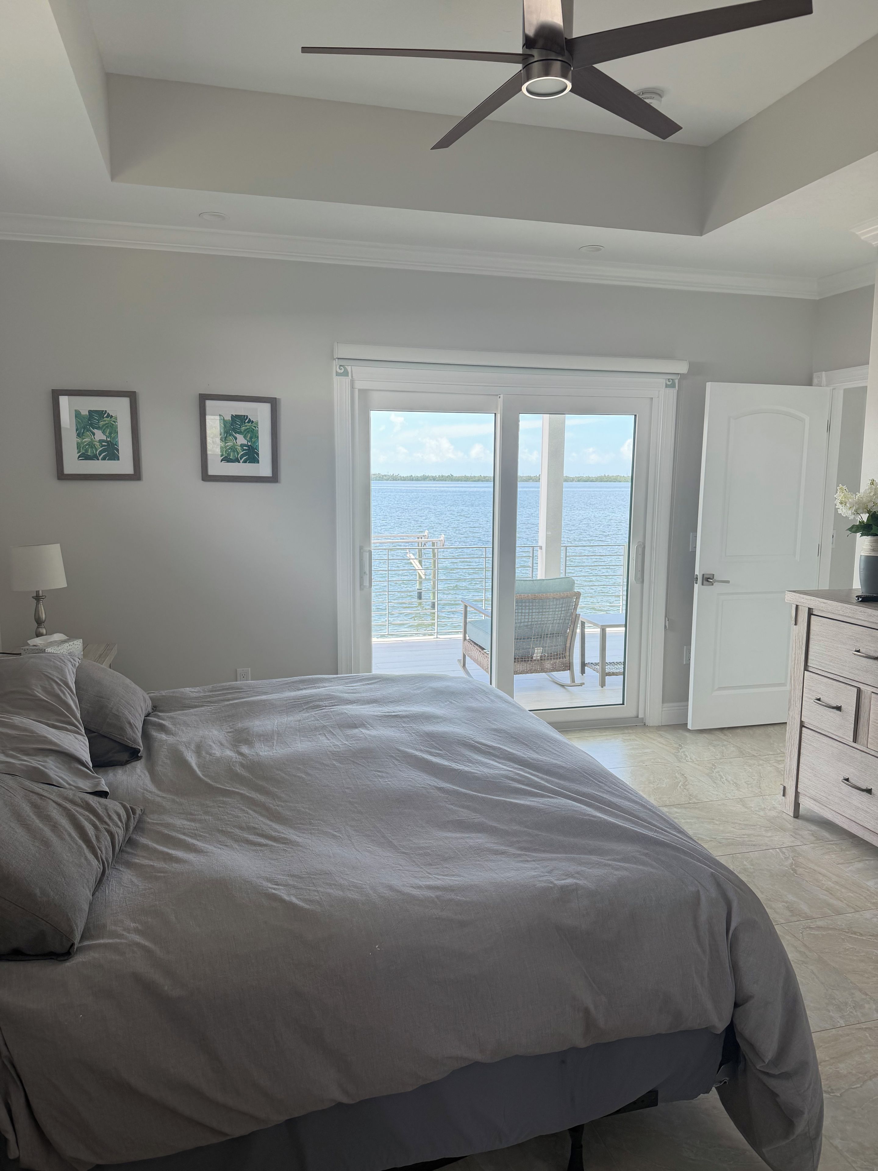 A bedroom with a large bed, a ceiling fan, a view of the water through the door, and a white dresser.