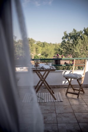 Une table en bois, sur laquelle se trouvent un livre, une tasse et un bocal de biscuits, est placée sur un balcon agrémenté d’une chaise et offrant une vue sur la verdure.