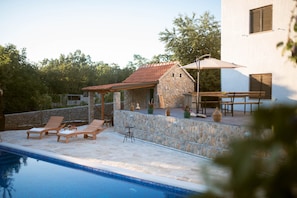 Un espace aménagé au bord de la piscine, avec des chaises longues, un mur en pierre et un bâtiment doté d’un toit en tuiles.
