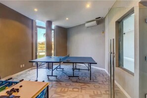 A room with a ping pong table, a foosball table, and a large window providing a view of the outdoors.