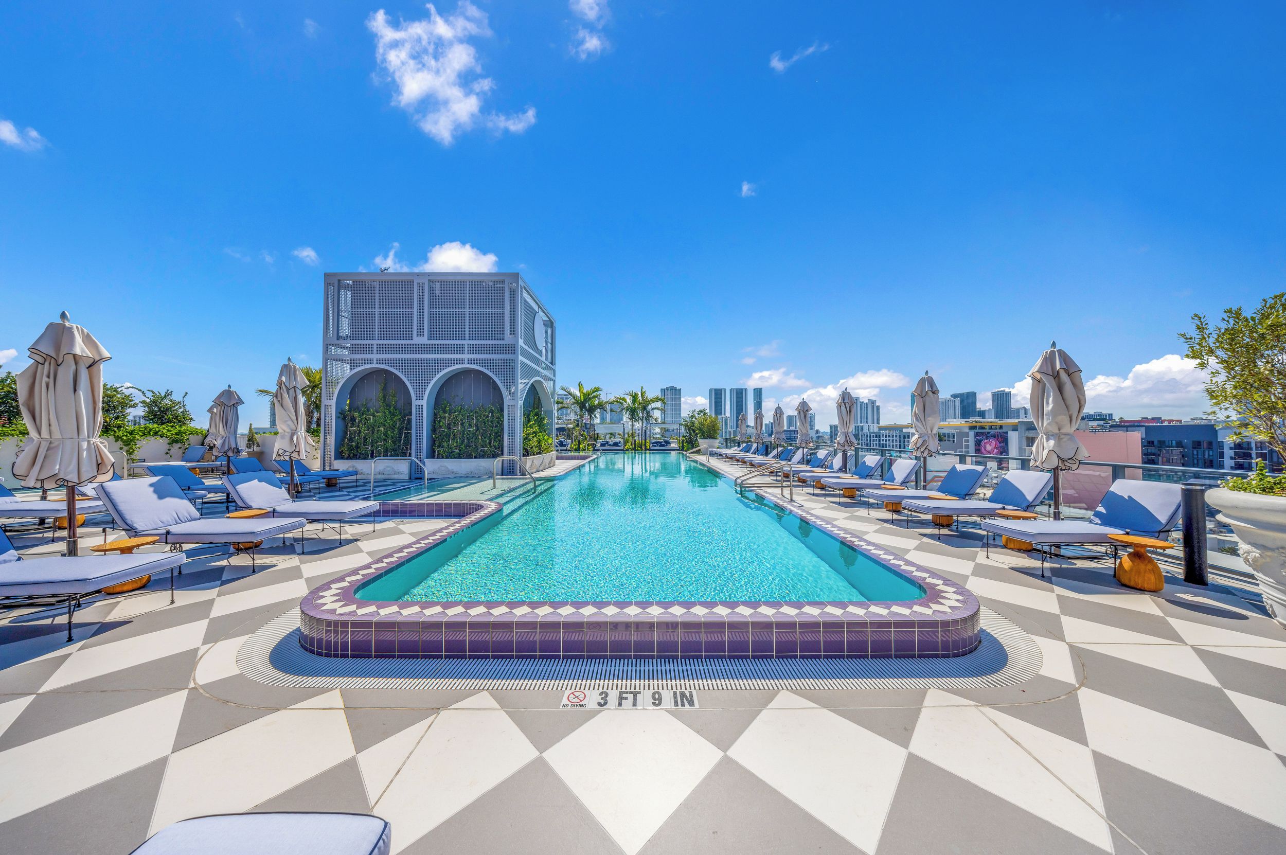 A rooftop pool with lounge chairs and umbrellas, a modern building with arched windows, and a clear blue sky.