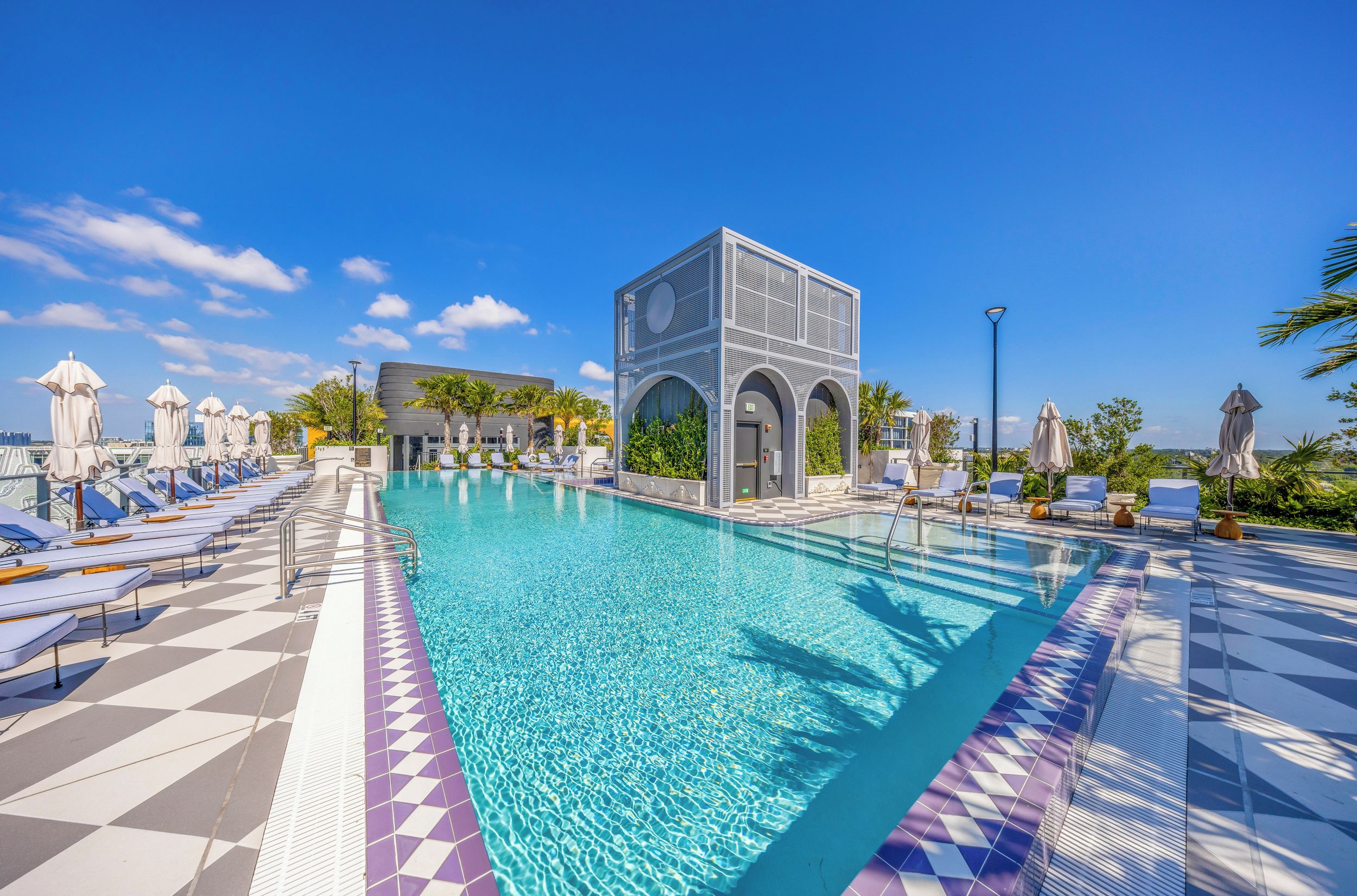 Piscina com espreguiçadeiras e guarda-sóis, cercada por palmeiras e um edifício moderno com fachada geométrica.