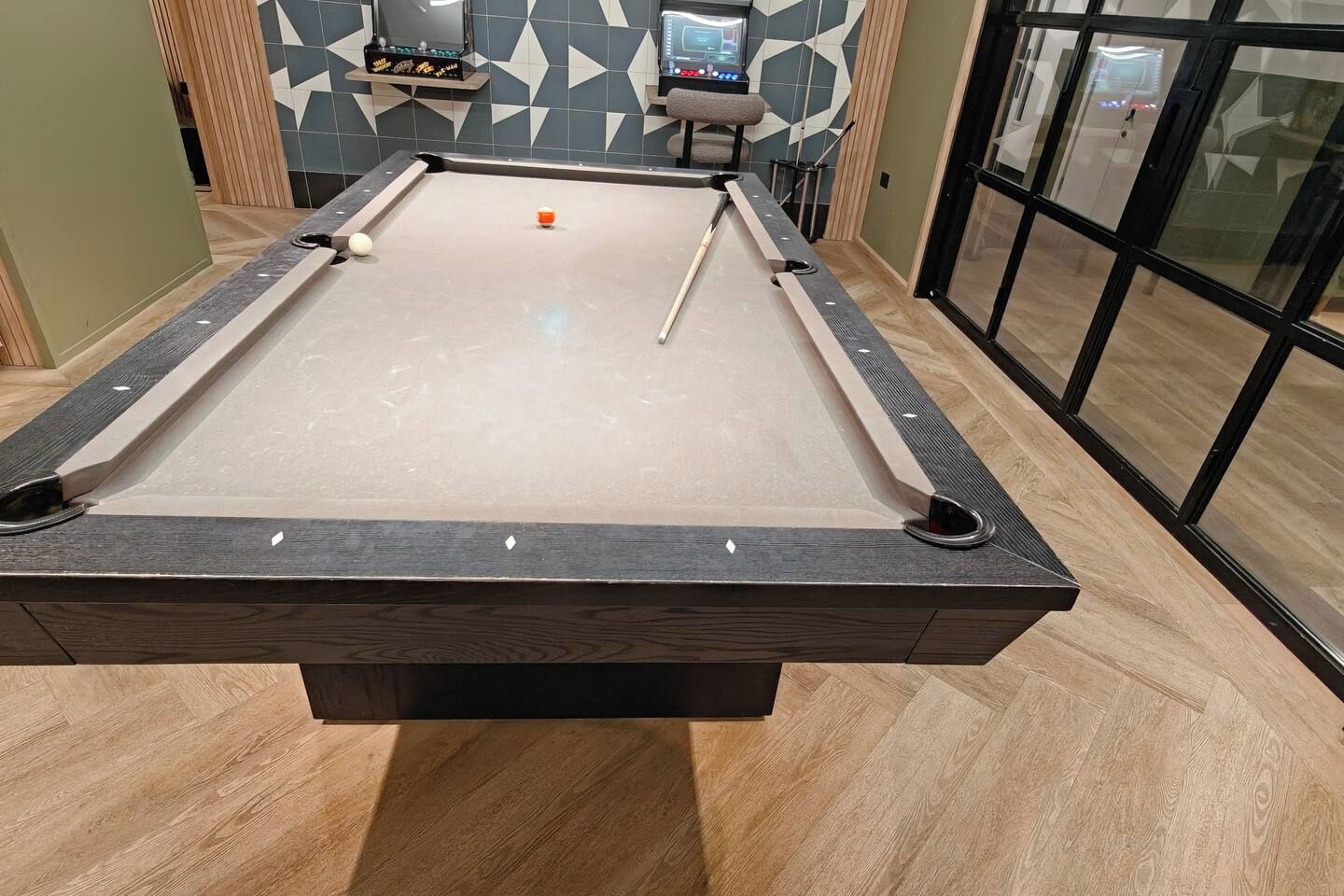 A pool table with a cue ball and an orange ball, surrounded by a modern room with geometric patterned walls and a glass door.