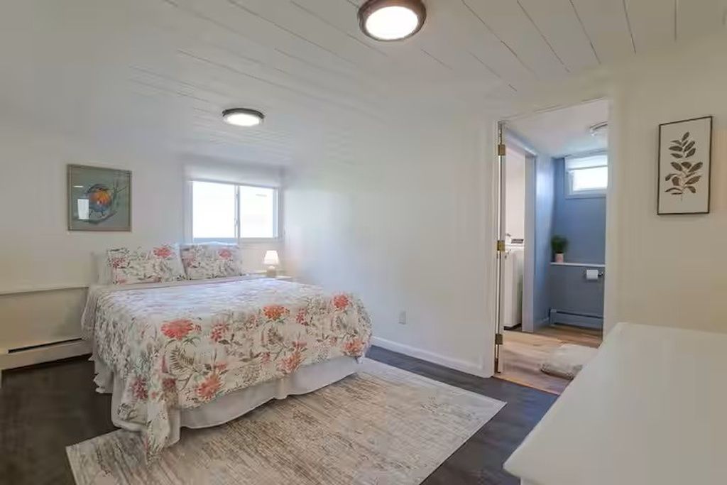 A bedroom with a bed, a window, a door leading to another room, and a wall-mounted picture.